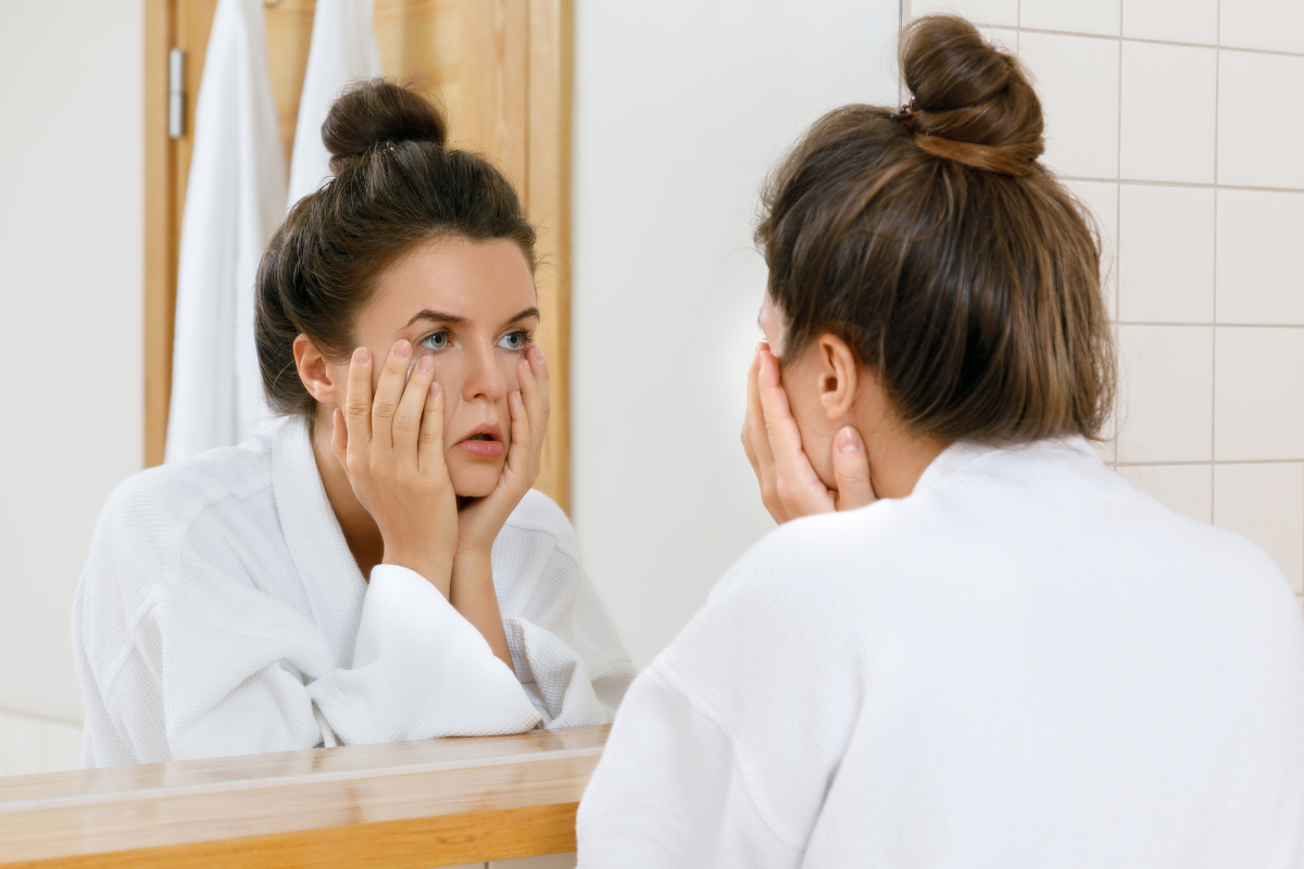 Mujer mirándose al espejo con expresión de cansancio mientras evalúa los cambios en su rostro más allá de las ojeras.